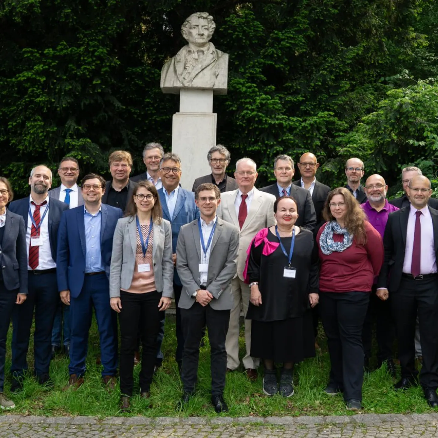 Eine Gruppe von Menschen steht vor einer Büste von Paul Johann Anselm von Feuerbach im Freien. Die Personen tragen formelle Kleidung und lächeln in die Kamera. Im Hintergrund sind grüne Büsche und Bäume zu sehen.