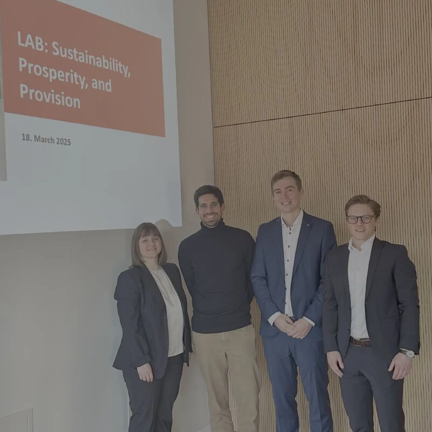 Vier Personen stehen lächelnd vor einer Präsentationsfolie mit der Aufschrift "LAB: Sustainability, Prosperity, and Provision, 18. March 2025". Sie befinden sich in einem modernen Raum mit Holzvertäfelung.