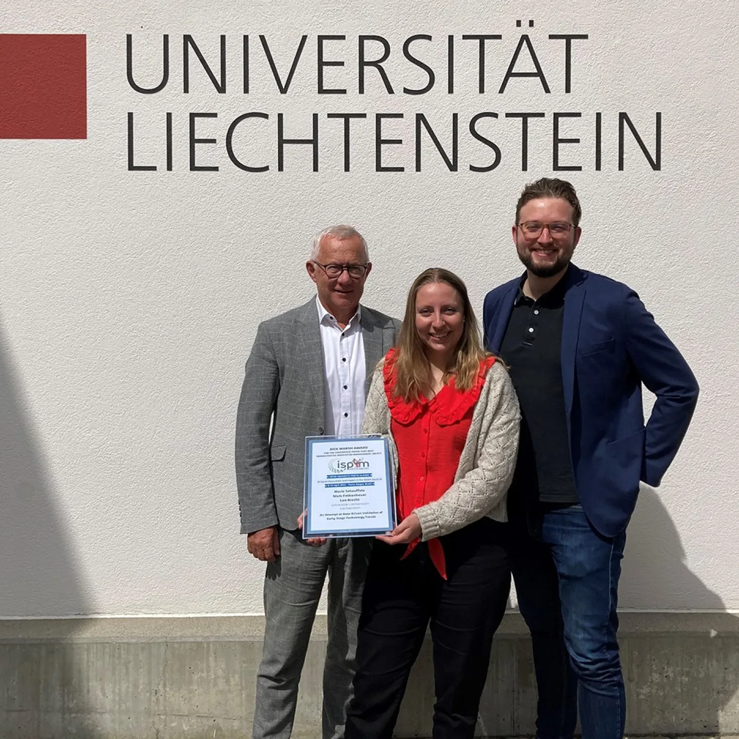 Drei Personen stehen vor einer Wand mit dem Logo der Universität Liechtenstein. Eine Person hält eine Auszeichnung in den Händen.