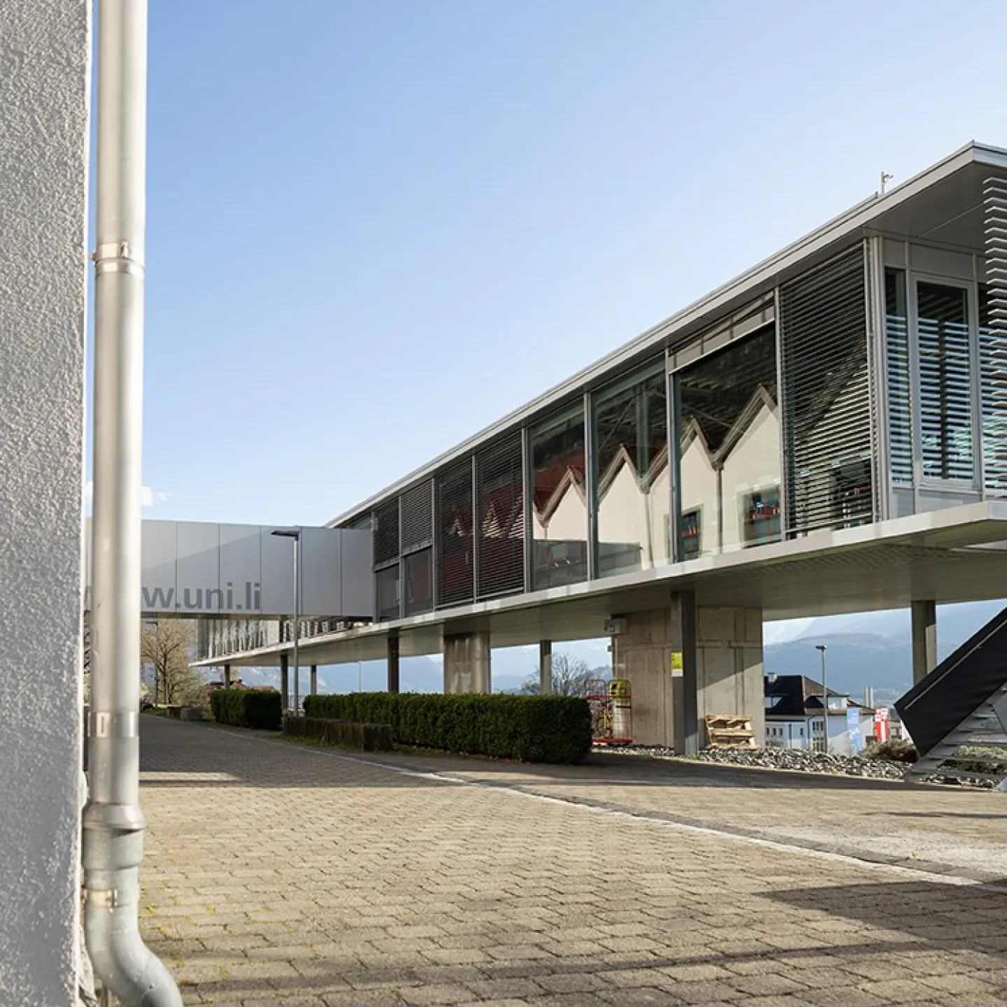 Modernes Gebäude der Universität Liechtenstein mit verglaster Fassade und Außentreppe, umgeben von einer gepflasterten Fläche und Hecken, vor einem klaren blauen Himmel.