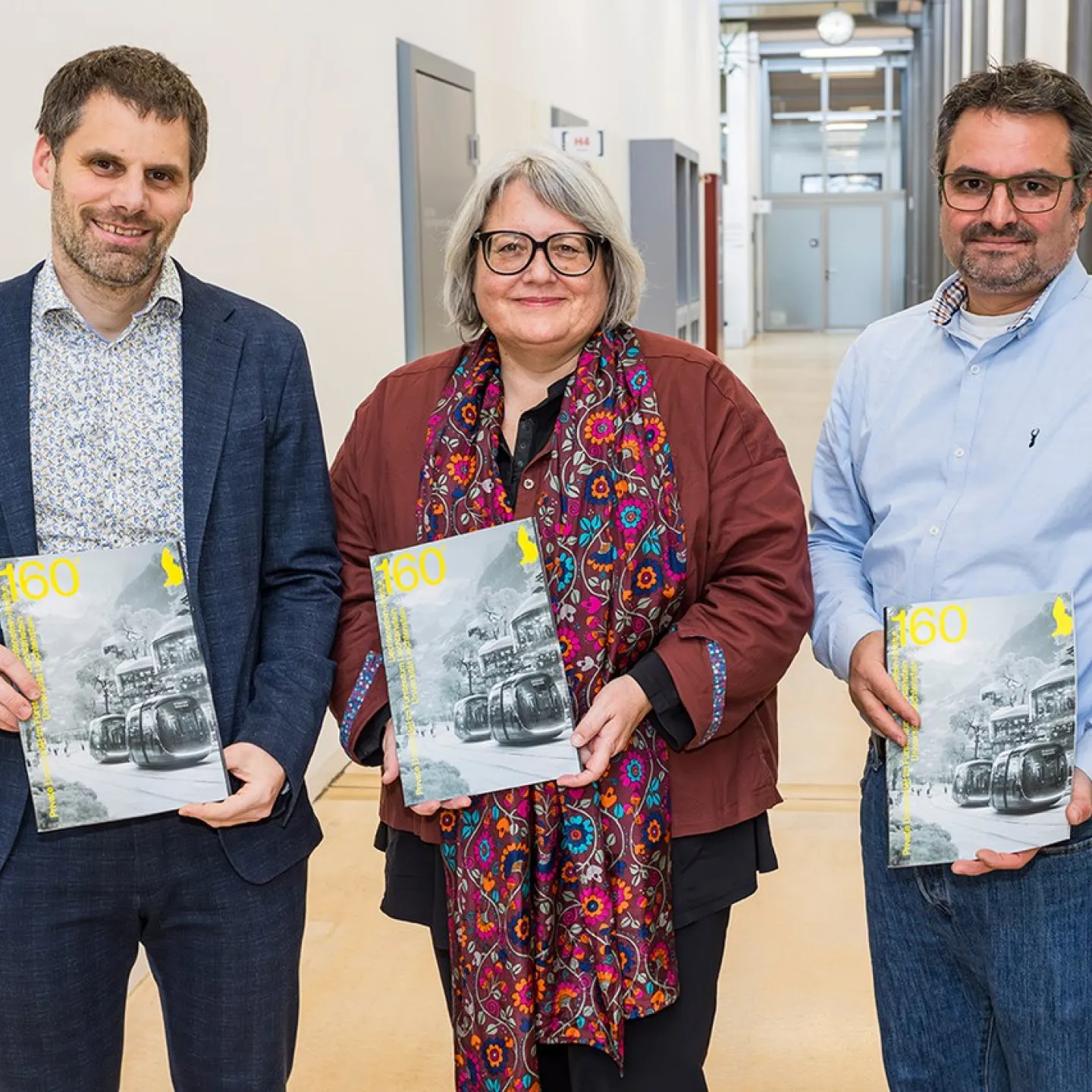 Drei Personen stehen in einem Flur und halten das Forschungsmagazin "160²" in den Händen. Das Magazin behandelt das Thema Künstliche Intelligenz.