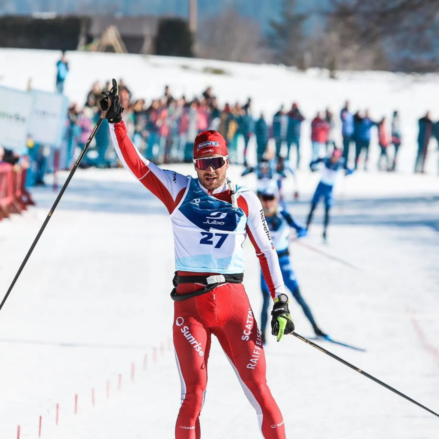 Langläufer überquert bei sonnigem Winterwetter als Sieger die Ziellinie eines Langlaufrennens, trägt einen rot-weißen Rennanzug mit Startnummer 27, hebt den Arm zum Jubel – im Hintergrund jubelnde Zuschauer und schneebedeckte Landschaft.