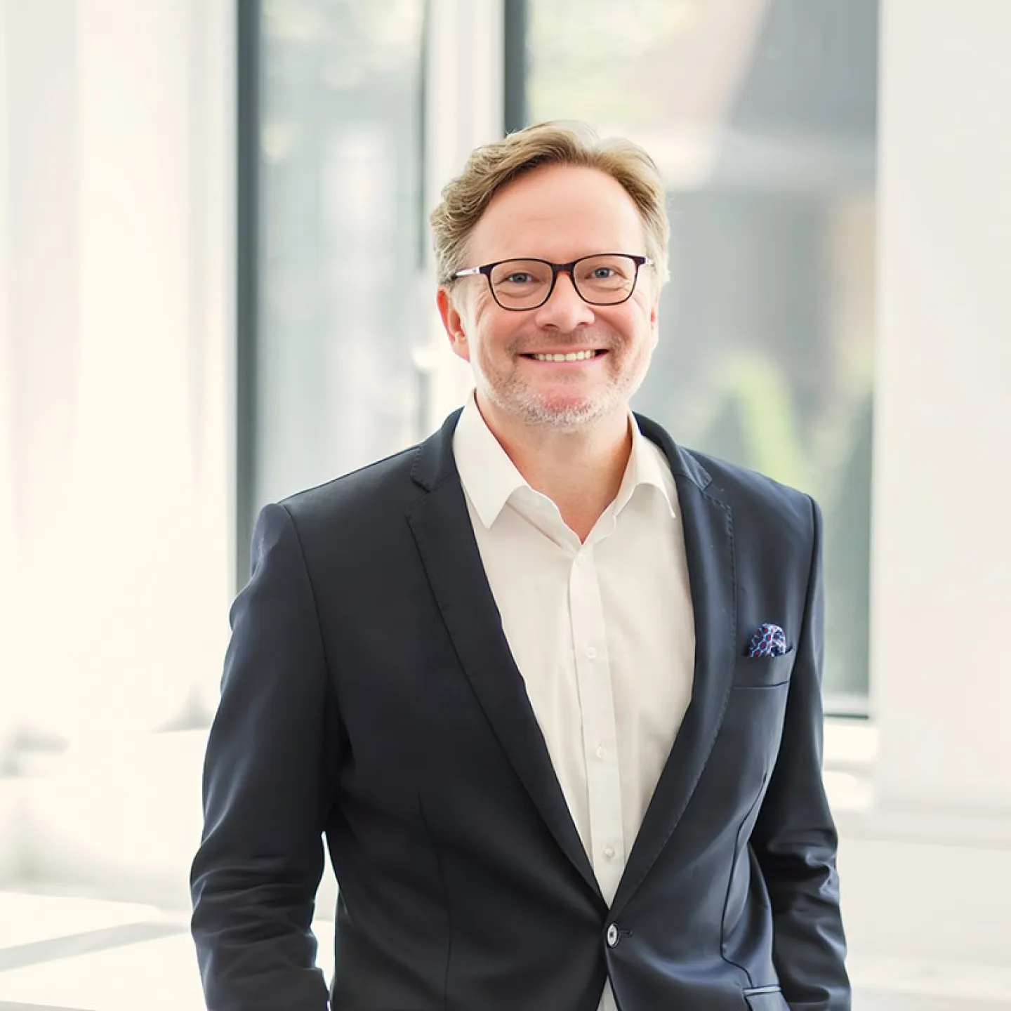 Ein lächelnder Mann in einem Anzug steht vor einem hellen Fenster.