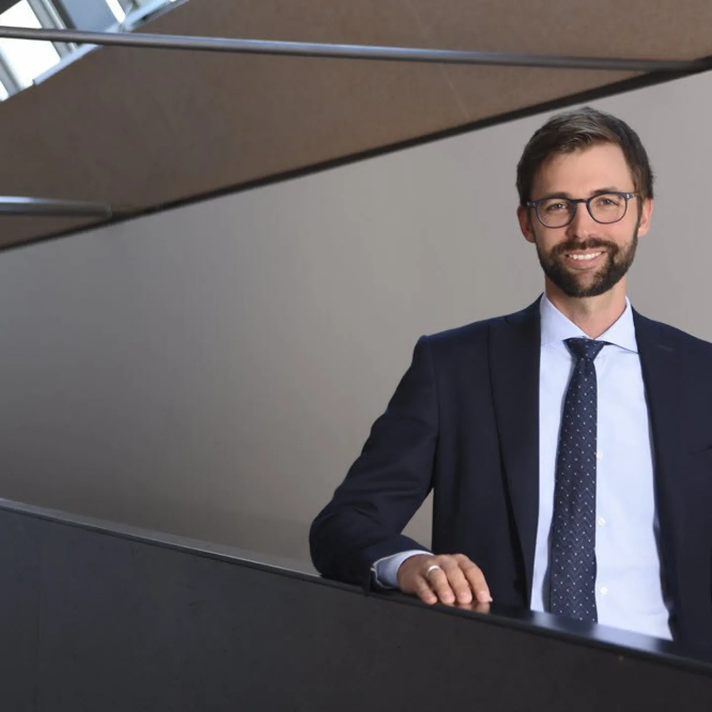 Ein Mann in Anzug und Krawatte steht lächelnd in einem modernen Gebäude mit großen Fenstern.