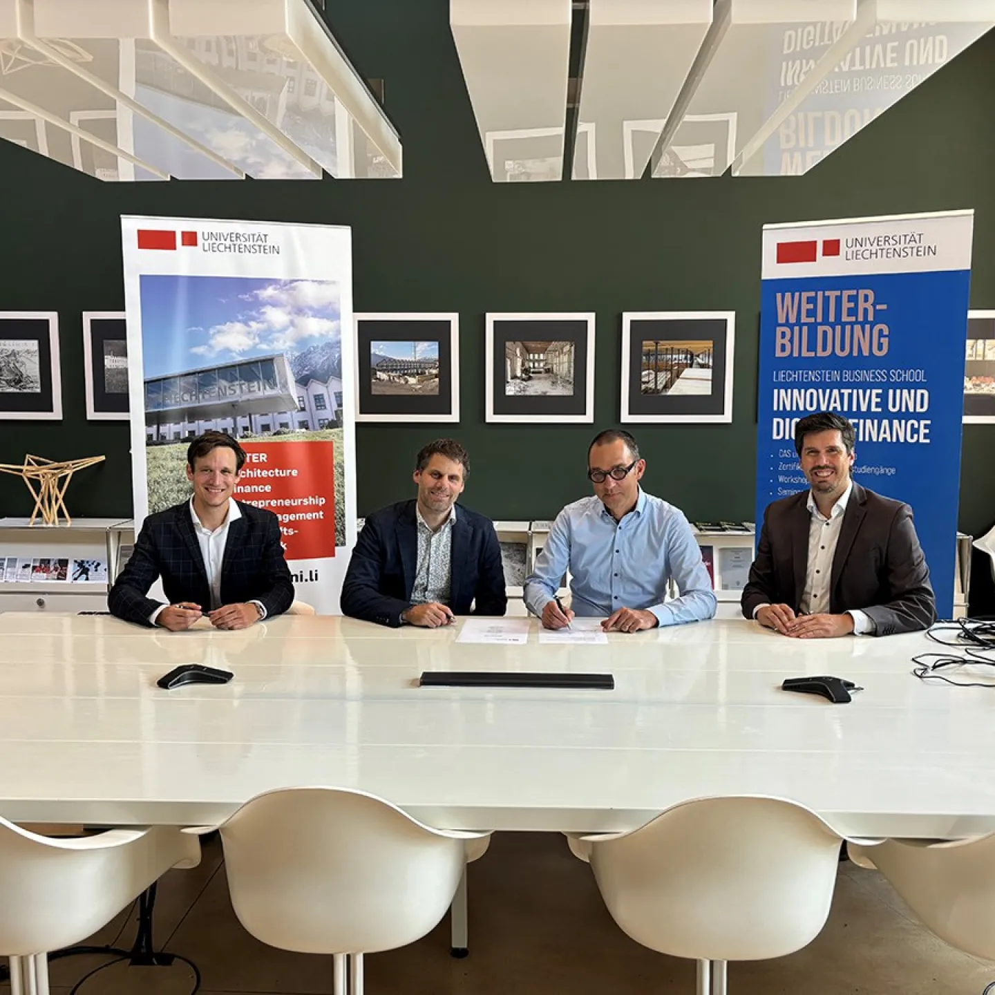 Vier Männer sitzen an einem Tisch in einem modernen Konferenzraum der Universität Liechtenstein. Im Hintergrund sind Banner mit dem Logo der Universität und Informationen zur Weiterbildung sichtbar.