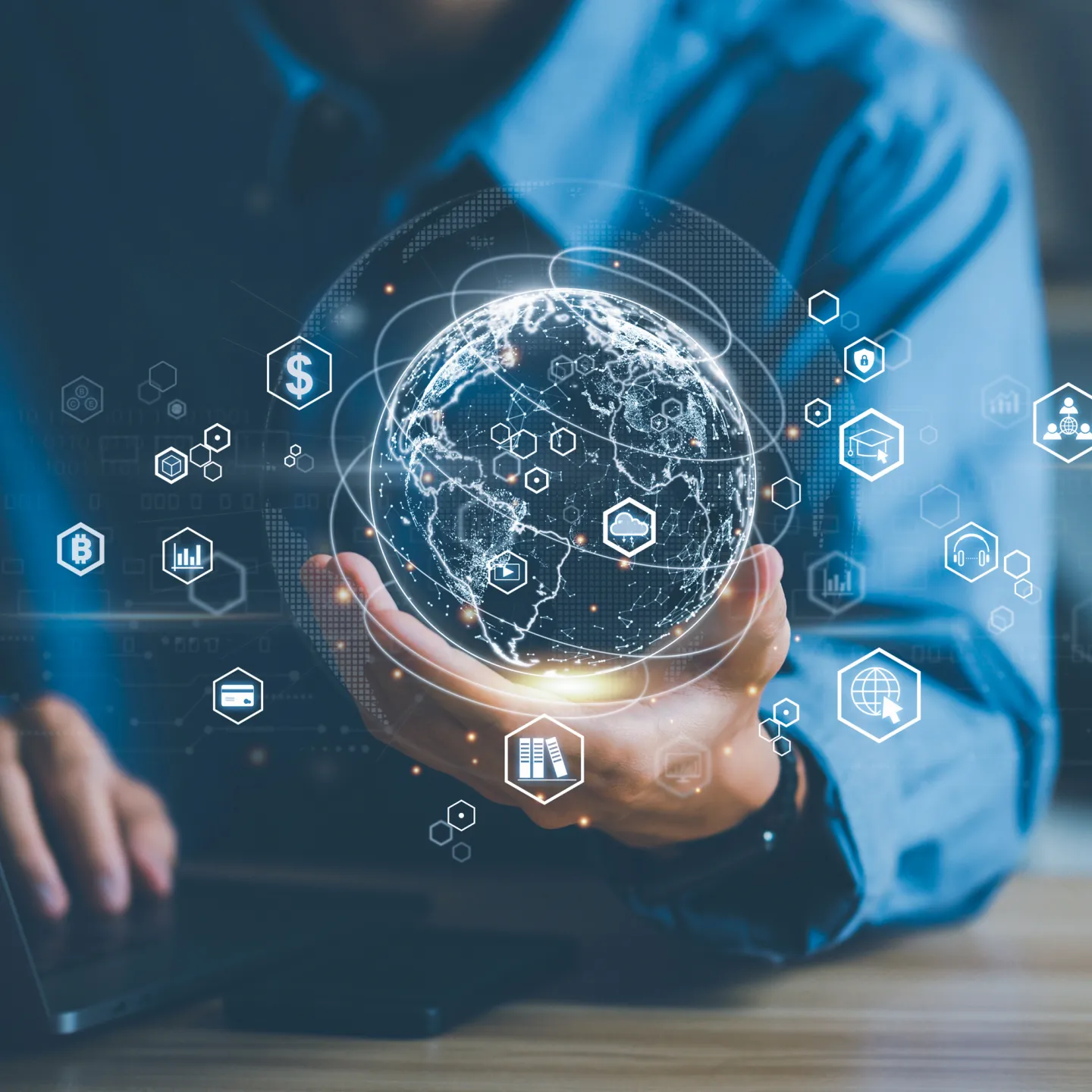 Person hält digitale Weltkugel mit globalen Wirtschaftssymbolen vor Laptop.