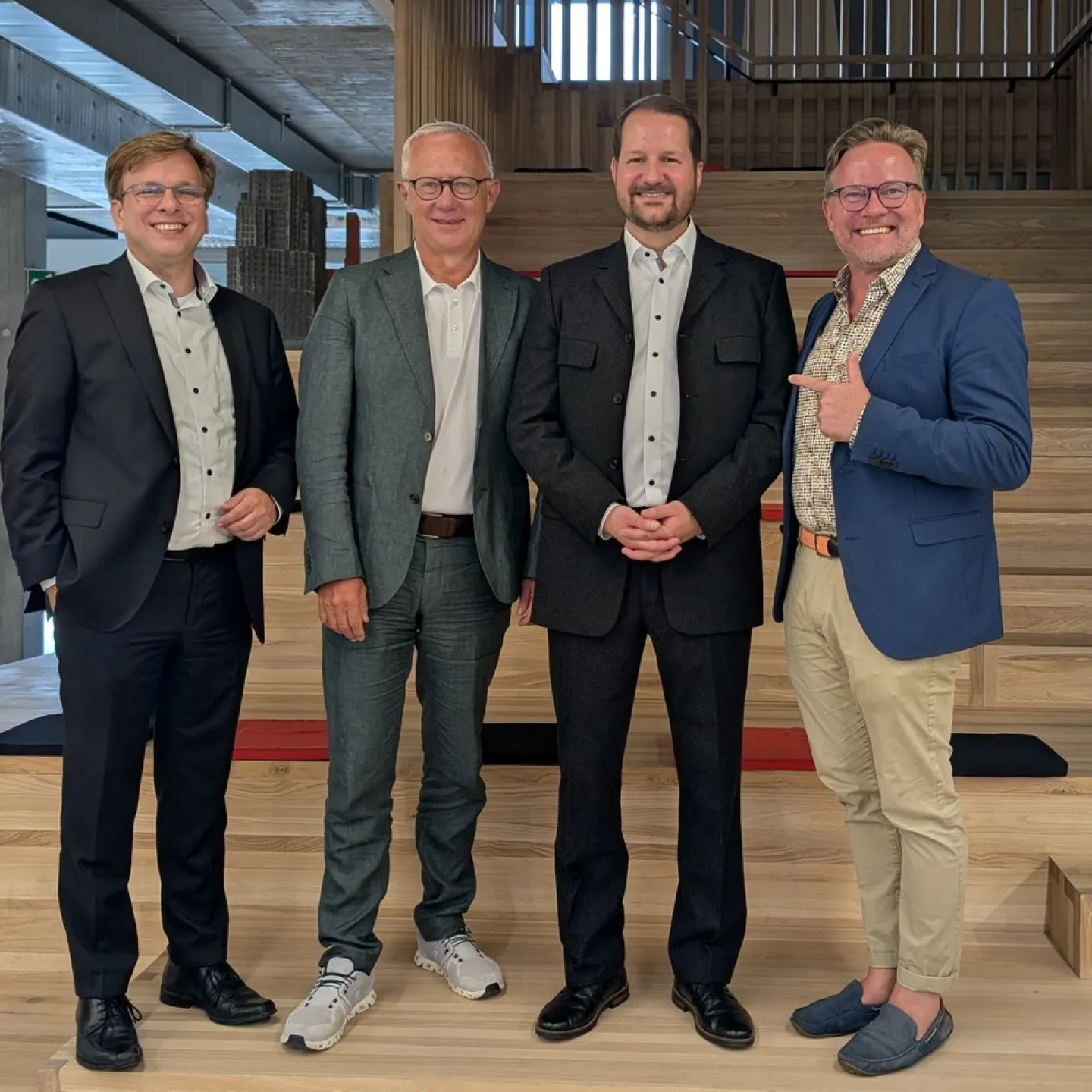 Vier Männer in formeller Kleidung stehen lächelnd auf einer Holztreppe in einem modernen Gebäude.