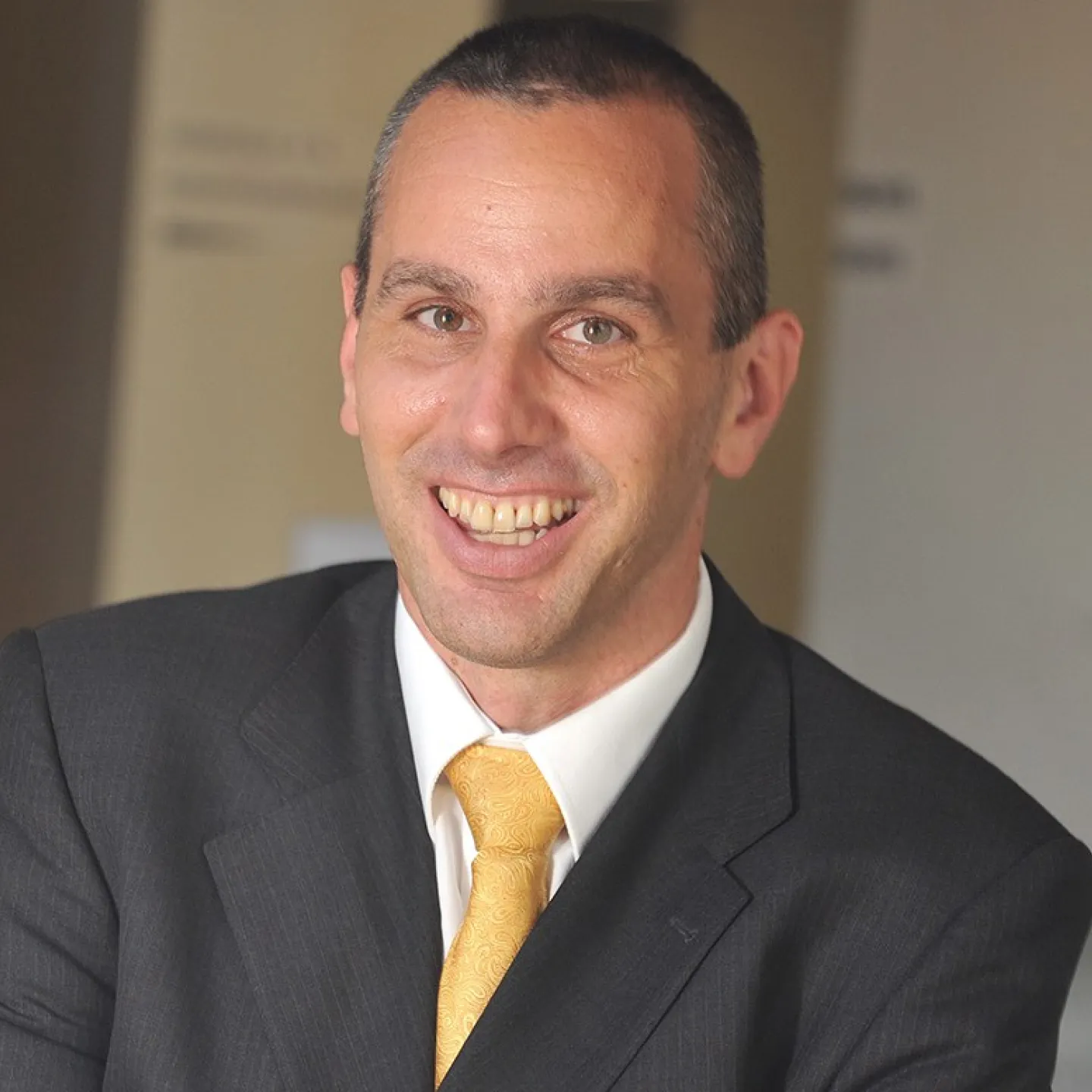 Porträt eines lächelnden Mannes in einem dunklen Anzug mit gelber Krawatte, vor einem neutralen Hintergrund.