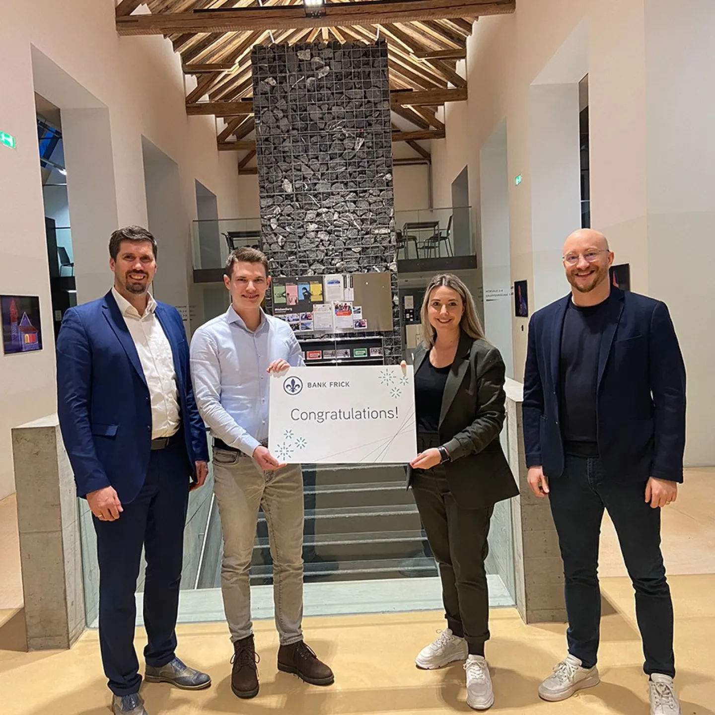 Vier Personen stehen in einem modernen Gebäude und halten ein Schild mit der Aufschrift "Congratulations!" von Bank Frick. Sie feiern die Vergabe eines Stipendiums für den Zertifikatsstudiengang Blockchain und FinTech an der Universität Liechtenstein.