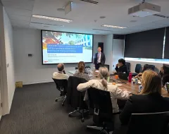 Ein Vortragender steht vor einer Präsentation mit dem Titel "Sustainable Taxation" in einem Konferenzraum. Mehrere Personen sitzen an einem Tisch und hören zu. Die Präsentation behandelt internationale Steuerpolitik und Nachhaltigkeit.