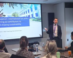 Ein Vortragender steht vor einer Präsentation mit dem Titel "Sustainable Taxation: International tax policy and reporting as contributors to achieving and shaping sustainability". Im Raum sitzen mehrere Personen, die dem Vortrag folgen.