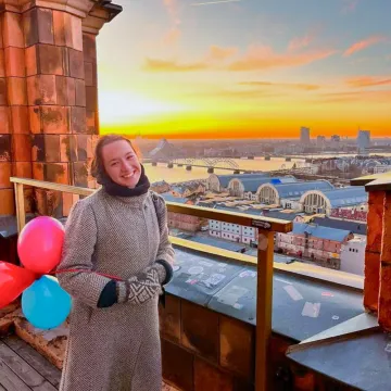 Studentin der Universität Liechtenstein auf einer Dachterrasse mit Blick über Riga bei Sonnenuntergang, umgeben von Luftballons.