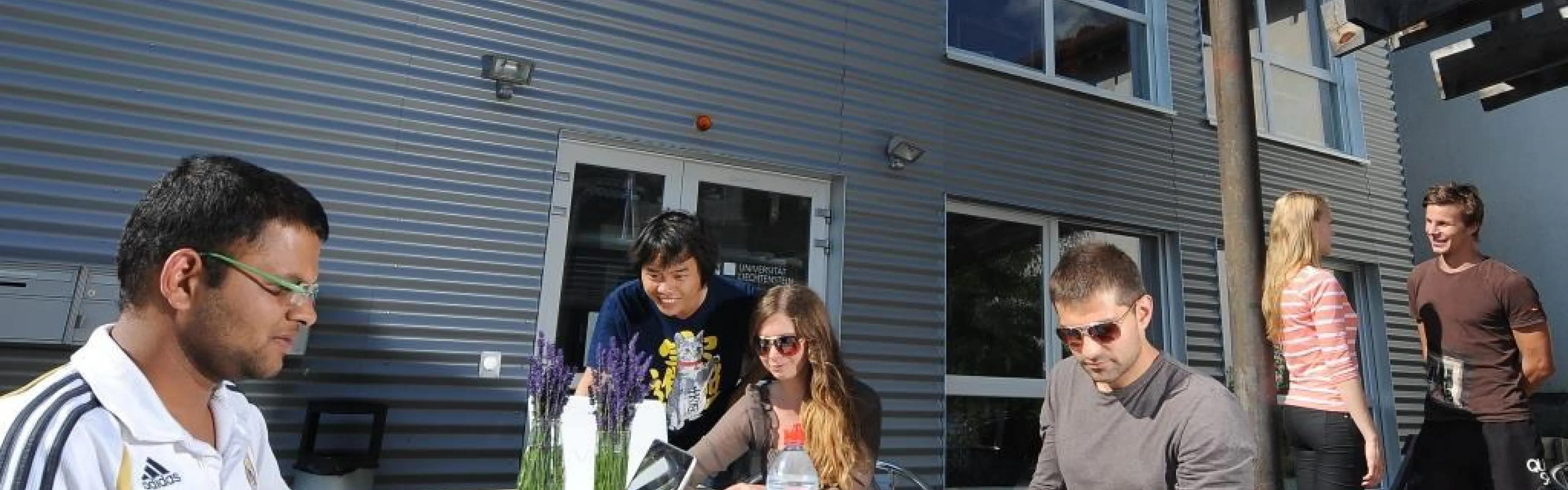 Eine Gruppe von Studierenden der Universität Liechtenstein sitzt und steht um einen Tisch vor der Uni Lichtenstein. Einige benutzen Laptops und Smartphones.