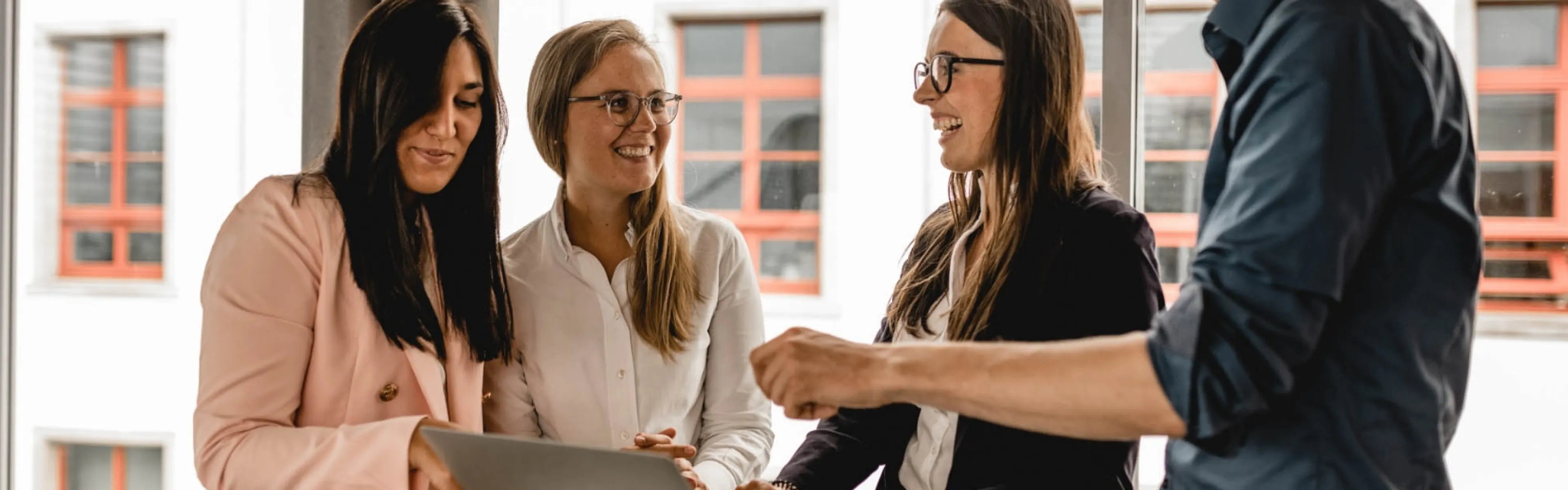 Eine Gruppe von vier Personen, drei Frauen und ein Mann, steht um einen Tisch mit einem Laptop und lacht gemeinsam. Sie tragen formelle Kleidung und scheinen an einem beruflichen Gespräch teilzunehmen.
