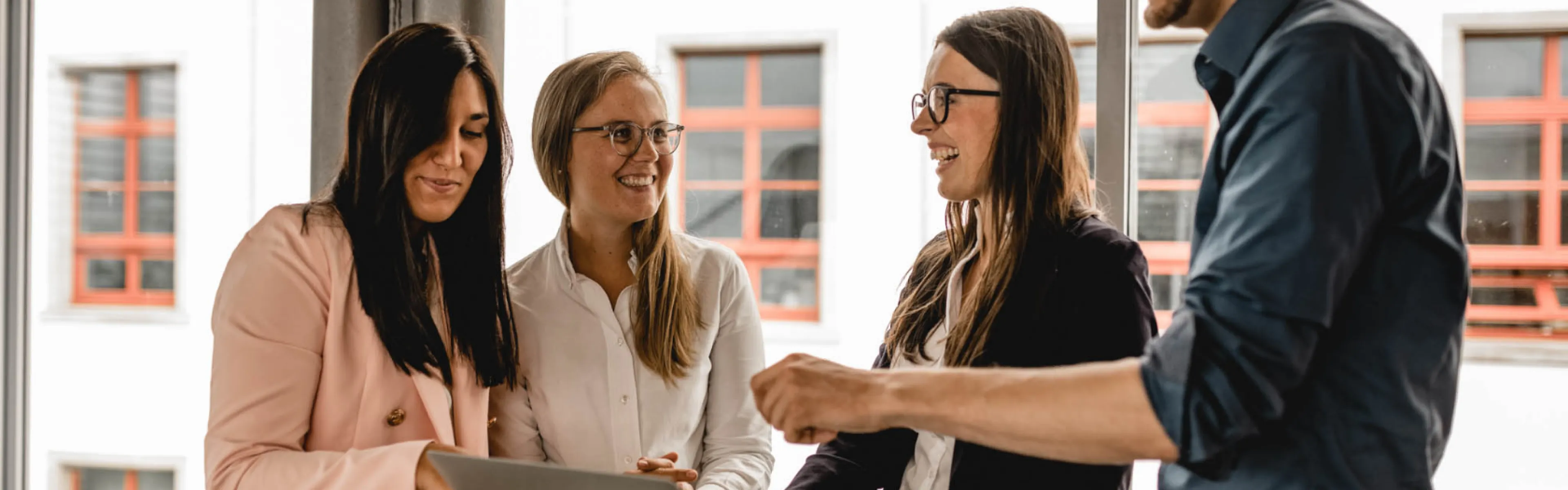 Vier Personen in formeller Kleidung arbeiten gemeinsam an einem Laptop, lächeln und diskutieren, während sie in einem modernen Büro stehen.
