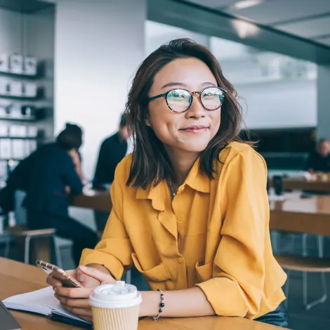 Eine junge Frau sitzt in der Mensa und blickt nachdenklich aus dem Fenster. Vor ihr liegen ein aufgeklappter Laptop, ein geöffnetes Notizbuch sowie ein Kaffee im To-Go-Becher, die ihre konzentrierte Arbeitsatmosphäre unterstreichen.
