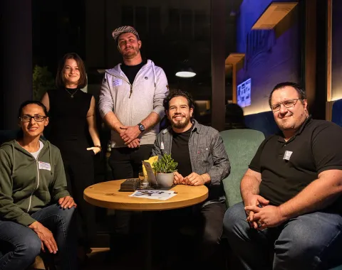 Eine Gruppe von fünf Personen sitzt und steht um einen Tisch in einem gemütlichen Café. Sie lächeln in die Kamera und wirken entspannt. Auf dem Tisch liegen Zeitschriften und eine Pflanze. Die Szene ist warm beleuchtet, was eine einladende Atmosphäre schafft.