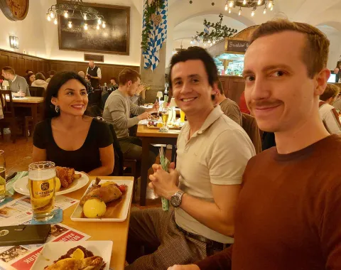 Drei Personen sitzen an einem Tisch in einem Restaurant und lächeln. Auf dem Tisch stehen traditionelle bayerische Speisen und Getränke. Im Hintergrund sind weitere Gäste und eine gemütliche Atmosphäre zu sehen.