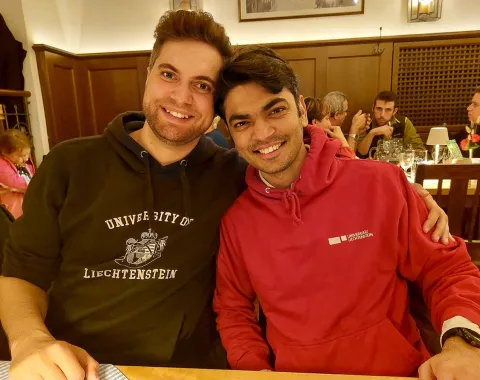 Zwei Personen sitzen lächelnd in einem Restaurant. Beide tragen Kapuzenpullover, einer mit dem Logo der Universität Liechtenstein. Im Hintergrund sind weitere Gäste an Tischen zu sehen. Die Atmosphäre wirkt gemütlich und gesellig.