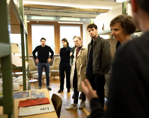 Eine Gruppe von Menschen steht in einem Raum mit Regalen und Möbeln. Sie hören einer Person zu, die außerhalb des Bildes spricht. Die Szene wirkt wie ein informelles Treffen oder eine Führung.