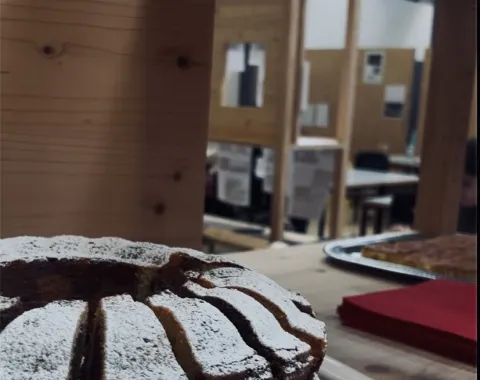 Ein frisch gebackener, mit Puderzucker bestäubter Kuchen steht auf einem Holztisch. Im Hintergrund sind Holzkonstruktionen und Tische in einem Raum zu sehen, die eine kreative und einladende Atmosphäre vermitteln.