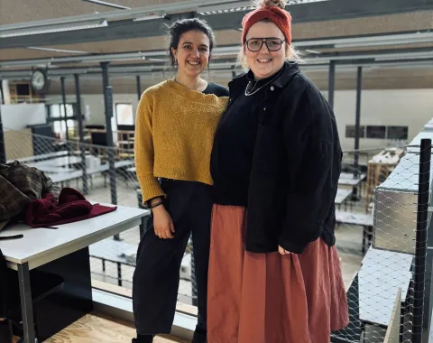 Zwei Personen stehen lächelnd in einem modernen Architekturatelier mit großen Fenstern und industriellem Design. Sie tragen lässige, kreative Kleidung. Im Hintergrund sind Arbeitsplätze und eine Uhr sichtbar.