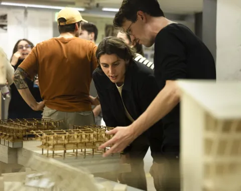 Zwei Personen betrachten aufmerksam Architekturmodelle in einem Ausstellungsraum. Im Hintergrund sind weitere Personen in Gespräche vertieft. Die Szene vermittelt eine Atmosphäre von Interesse und Austausch im Rahmen eines universitären Architektur-Events.
