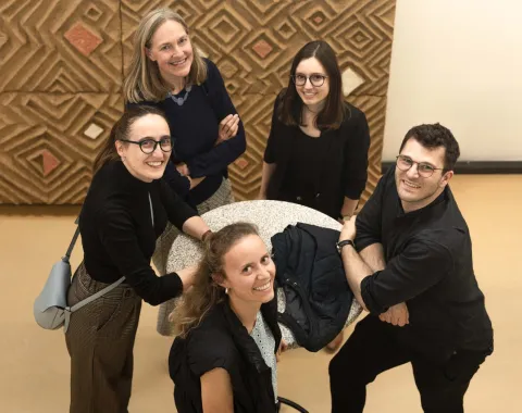 Eine Gruppe von fünf Personen steht um einen runden Tisch und lächelt in die Kamera. Der Hintergrund zeigt ein geometrisches Muster an der Wand. Die Szene wirkt freundlich und einladend.