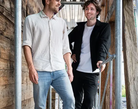 Zwei junge Männer stehen lächelnd auf einem Gerüst in einem Holzbau. Sie tragen legere Kleidung und wirken entspannt. Die Umgebung deutet auf eine Baustelle oder ein architektonisches Projekt hin.