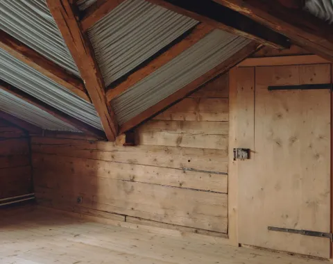 Innenansicht eines Dachbodens mit Holzwänden und Holzboden, sichtbare Dachbalken und Wellblechverkleidung. Eine Holztür befindet sich an der rechten Wand.