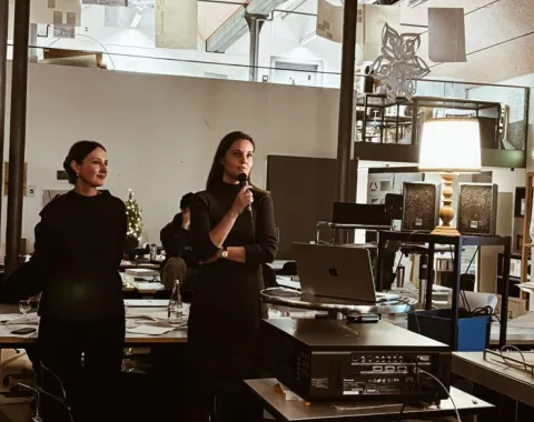 Zwei Personen stehen in einem modernen Architekturstudio. Eine Person hält ein Mikrofon und spricht, während die andere zuhört. Im Hintergrund sind Arbeitsplätze und Architekturmodelle zu sehen.