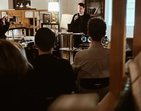 Ein Vortragender spricht vor einer Gruppe von Studierenden in einem Raum mit Präsentationstechnik. Im Hintergrund sind Regale und eine Lampe sichtbar. Die Atmosphäre wirkt konzentriert und lernorientiert.