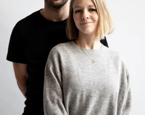 Ein Mann und eine Frau stehen vor einem neutralen Hintergrund. Beide blicken direkt in die Kamera. Die Frau trägt einen grauen Pullover, der Mann ein schwarzes T-Shirt.
