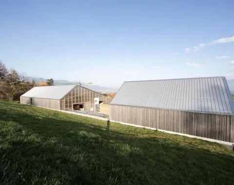 Zwei moderne Gebäude mit Holz- und Glasfassaden in einer ländlichen Umgebung. Die Architektur ist schlicht und kontextbezogen, eingebettet in eine grüne Landschaft.