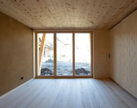 Innenansicht eines minimalistischen Raumes mit Holzverkleidung und großem Fenster, das den Blick auf eine ländliche Umgebung freigibt. Der Raum ist lichtdurchflutet und zeigt eine moderne, nachhaltige Architektur.