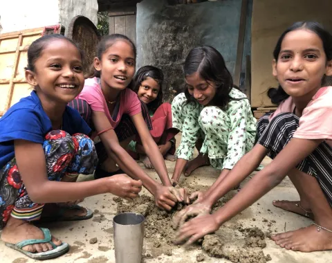 Eine Gruppe von lachenden Kindern kniet auf dem Boden und formt gemeinsam Ton oder Lehm. Die Szene vermittelt Freude und Zusammenarbeit in einem handwerklichen Kontext.