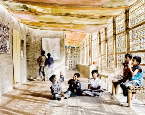 Ein heller Korridor mit Wänden aus natürlichen Materialien wie Lehm und Bambus. Kinder sitzen und stehen entspannt im Raum, der mit bunten Stoffen dekoriert ist. Die Architektur verbindet traditionelle Bauweisen mit modernem Design.