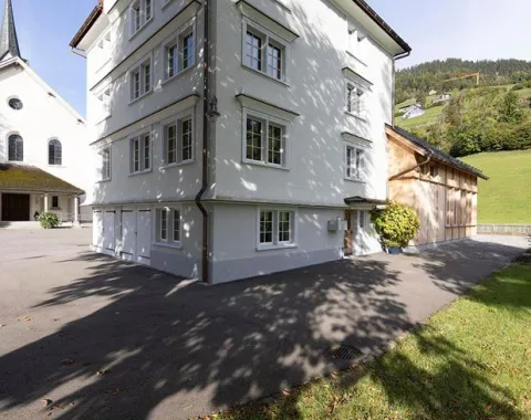 Ein weißes, mehrstöckiges Gebäude mit traditionellen Fenstern steht neben einer Kirche im ländlichen St. Galler Rheintal. Im Hintergrund sind grüne Hügel zu sehen. Der Schatten eines Baumes fällt auf den gepflasterten Vorplatz.