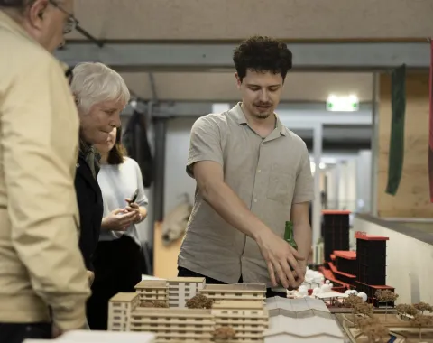 Eine Gruppe von Menschen betrachtet ein Architekturmodell in einem Innenraum. Ein Mann zeigt auf das Modell und erklärt es den anderen Anwesenden. Im Hintergrund sind weitere Modelle und Regale zu sehen.