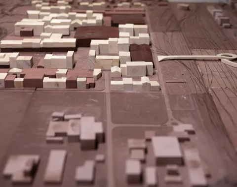 Ein Architekturmodell zeigt eine städtische Umgebung mit Gebäuden in verschiedenen Größen und Formen. Die Struktur besteht aus hellen und dunklen Holzblöcken, die Straßen und Freiflächen umgeben. Das Modell veranschaulicht städtebauliche Planung und Design.