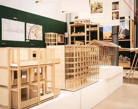 Architekturmodelle aus Holz in einem Ausstellungsraum der Universität Liechtenstein. Die Modelle zeigen detaillierte Gebäudestrukturen und sind Teil der Präsentation der Liechtenstein School of Architecture. Im Hintergrund sind Pläne und weitere Modelle sichtbar.