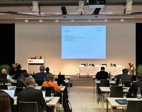 Ein Konferenzraum mit Teilnehmern, die an Tischen sitzen und einem Vortrag folgen. Eine Person steht am Rednerpult vor einer Leinwand, auf der eine Präsentation mit dem Titel "Agenda" angezeigt wird. Der Raum ist modern eingerichtet, mit mehreren Tischen und Stühlen.