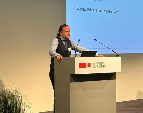 Ein Redner steht an einem Podium mit dem Logo der Universität Liechtenstein. Im Hintergrund ist eine Präsentationsfolie mit dem Titel "Reflexion" und den Fragen "Wo stehen wir?", "Was betrachten wir?" und "Welche Erkenntnisse erhalten wir?" zu sehen. Links im Bild sind Pflanzen dekorativ platziert.