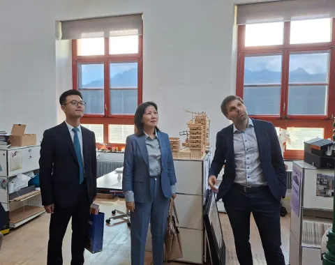 Drei Personen stehen in einem Büro mit großen Fenstern, durch die Berge sichtbar sind. Sie betrachten interessiert etwas außerhalb des Bildes. Im Hintergrund sind Regale und Modelle zu sehen.