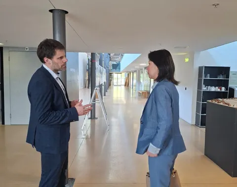 Zwei Personen in einem modernen Gebäude im Gespräch. Beide tragen Business-Kleidung. Im Hintergrund sind helle Flure und moderne Einrichtungselemente zu sehen.