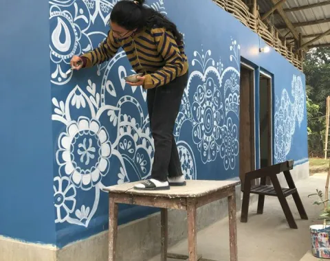 Eine Person bemalt die blaue Außenwand einer Schule in Indien mit weißen, floralen Mustern. Sie steht auf einem Holztisch, um die oberen Bereiche zu erreichen. Das Gebäude hat ein Wellblechdach und ist Teil eines Bildungsprojekts.