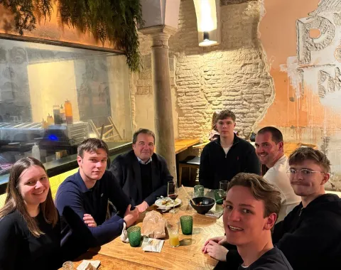 Eine Gruppe von sieben Personen sitzt an einem Tisch in einem Restaurant. Sie lächeln in die Kamera. Im Hintergrund sind eine gemauerte Wand und Pflanzen zu sehen.