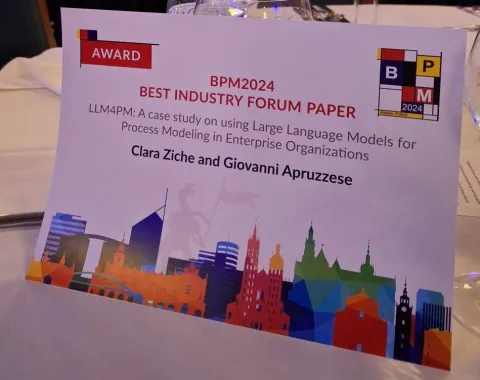 Ein Tischaufsteller mit der Aufschrift "BPM2024 Best Industry Forum Paper" und dem Titel der ausgezeichneten Masterarbeit von Clara Ziche und Giovanni Apruzzese. Im Hintergrund sind stilisierte Skyline-Elemente zu sehen.