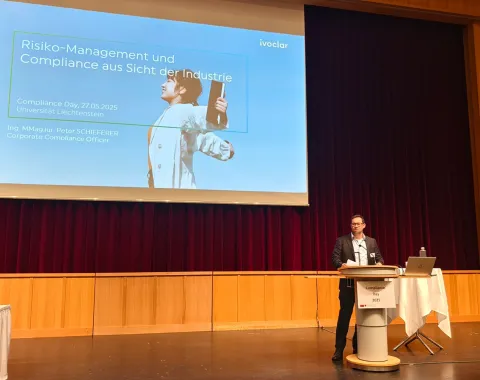 Ein Redner steht auf einer Bühne vor einem großen Bildschirm, der eine Präsentation mit dem Titel „Risiko-Management und Compliance aus Sicht der Industrie“ zeigt. Der Raum hat Holzwände und einen roten Vorhang. Ein Tisch mit Laptop und Mikrofon steht neben dem Redner.