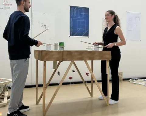 Zwei Personen stehen an einem hölzernen Tischmodell und diskutieren. Im Hintergrund sind Architekturzeichnungen an der Wand zu sehen. Die Szene spielt in einem Designstudio der Universität Liechtenstein.